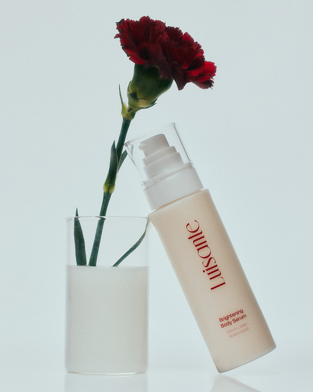 Luisante brightening body serum bottle beside a red carnation flower in clear glass with white liquid